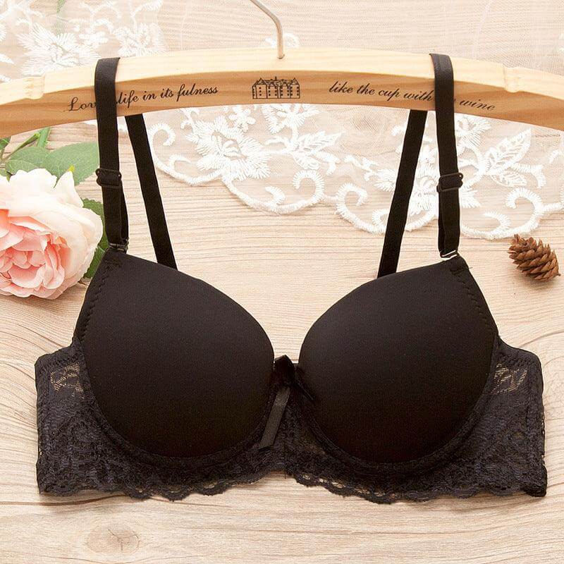 Black underwire gathered bra for small bust with lace and bow details on a wooden hanger, lace background, and floral decor.