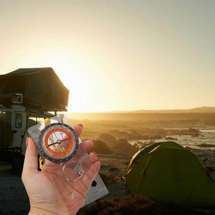 Hand holding 2-In-1 Compass With Map Ruler For Hiking And Outdoors against coastal campsite sunrise for trekking navigation