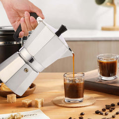 Pouring fresh espresso from RAINBEAN 3-Cup Stovetop Moka Pot into a glass cup on a wooden table with coffee beans and sugar cubes