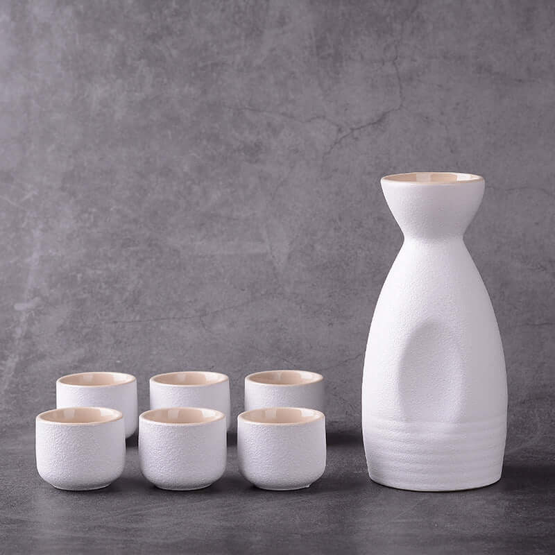 Japanese ceramic sake brewing ensemble with white sake bottle and six matching sake cups against gray background.