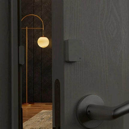 Xiaomi Intelligent Mini Door and Window Sensor for Home Security mounted on interior doorframe monitoring open door with lamp-lit room