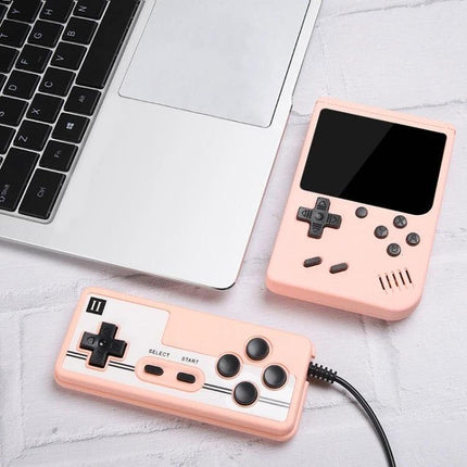 Macaron Mini Handheld Game Console with 800 Built-in Games - pink 3-inch LCD retro portable gaming device with wired controller next to laptop