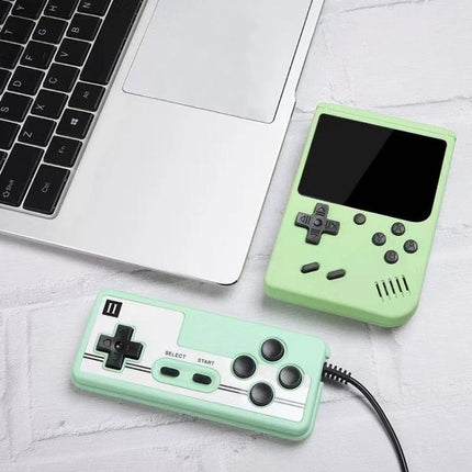 Macaron Mini Handheld Game Console with 800 Built-in Games in pastel green next to laptop and wired controller on white surface