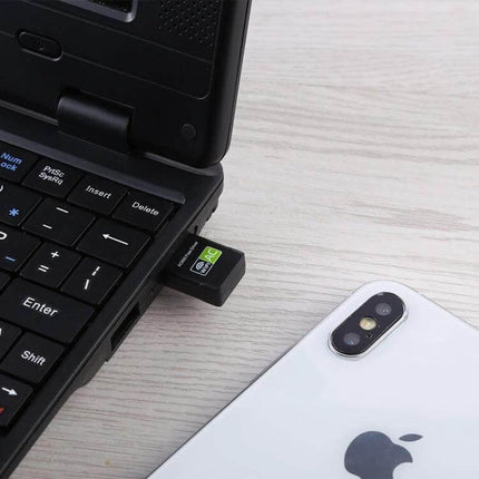 Dual Band USB WiFi Adapter for Wireless Internet Connectivity plugged into laptop USB port beside a smartphone, showing compact dual-band wireless dongle