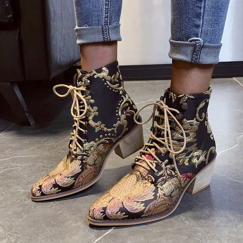 Stylish embroidered satin ankle boots with high chunky heels in black and gold, worn with cuffed jeans on a grey tiled floor.