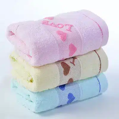 Caring Pure Cotton Face Towel for Gentle Daily Use 1 Three Neatly Stacked Towels In Pink, Yellow, And Blue With Heart Patterns And Text.