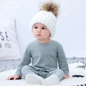 Toddler wearing a gray outfit and a white knitted hat with a pom-pom sitting on a soft surface.