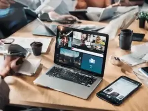A silver laptop displaying video calls and a smartphone on a wooden table among other devices.