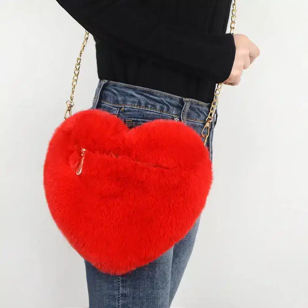Chic Plush Chain Shoulder Bag for Valentine's Day Delight 2 Red Heart-Shaped Handbag Made Of Fur With A Gold Chain Strap, Held By A Person.