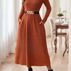 A woman wearing a rust orange ribbed midi dress with long sleeves and a black belt, standing indoors.