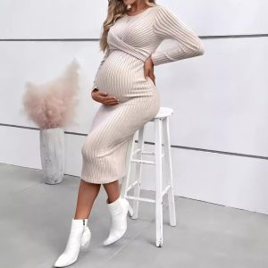 Woman wearing a beige ribbed maternity dress while sitting on a stool, holding her belly.