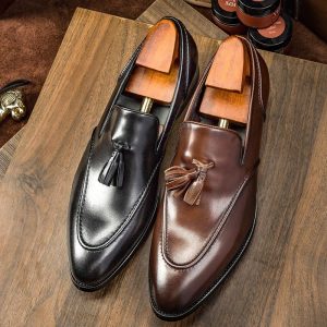 Polished black and brown leather tassel loafers with wooden shoe trees on a wooden surface, showcasing high-quality men's dress shoes for formal or business wear.