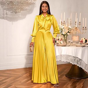 Woman wearing a yellow satin jumpsuit with a bow tie and pleated design, standing beside a table with candles.