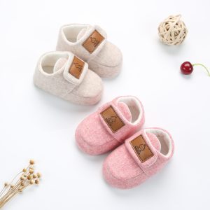 Soft baby slippers in pink and beige with brand labels, placed on a white background with cherry, wicker ball, and dried flowers for a cozy, stylish baby footwear display.