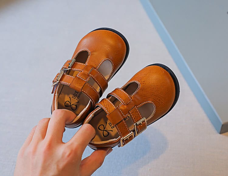 Princess Soft Sole Girls College Style Baby Shoes - Adorable & Comfortable 5 Hand Holding Children'S Brown Leather Shoes With Buckles On A Neutral Background.