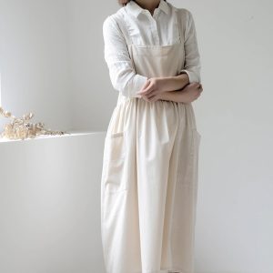 A person wearing a light beige apron dress over a white shirt, standing with arms crossed.