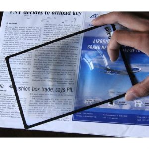 A hand holding a transparent plastic sheet with a black frame above a newspaper.
