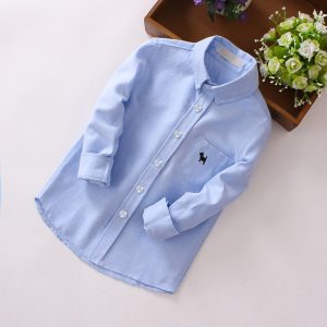 Soft Light Blue Boys' Formal Shirt With Front Buttons And A Small Black Dog Logo On Chest Pocket, Perfect For Children’s Business Or Casual Wear, Displayed On A Clean Surface With Decorative Flowers In The Background.