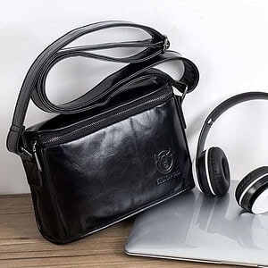 Stylish black leather briefcase with shoulder strap next to laptop and wireless headphones, ideal for business professionals and office work.