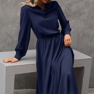 Elegant navy blue long-sleeve maxi dress with a cinched waist, displayed on a model sitting on a minimalist white table against a concrete background, perfect for fashion, formal wear, and stylish occasions.